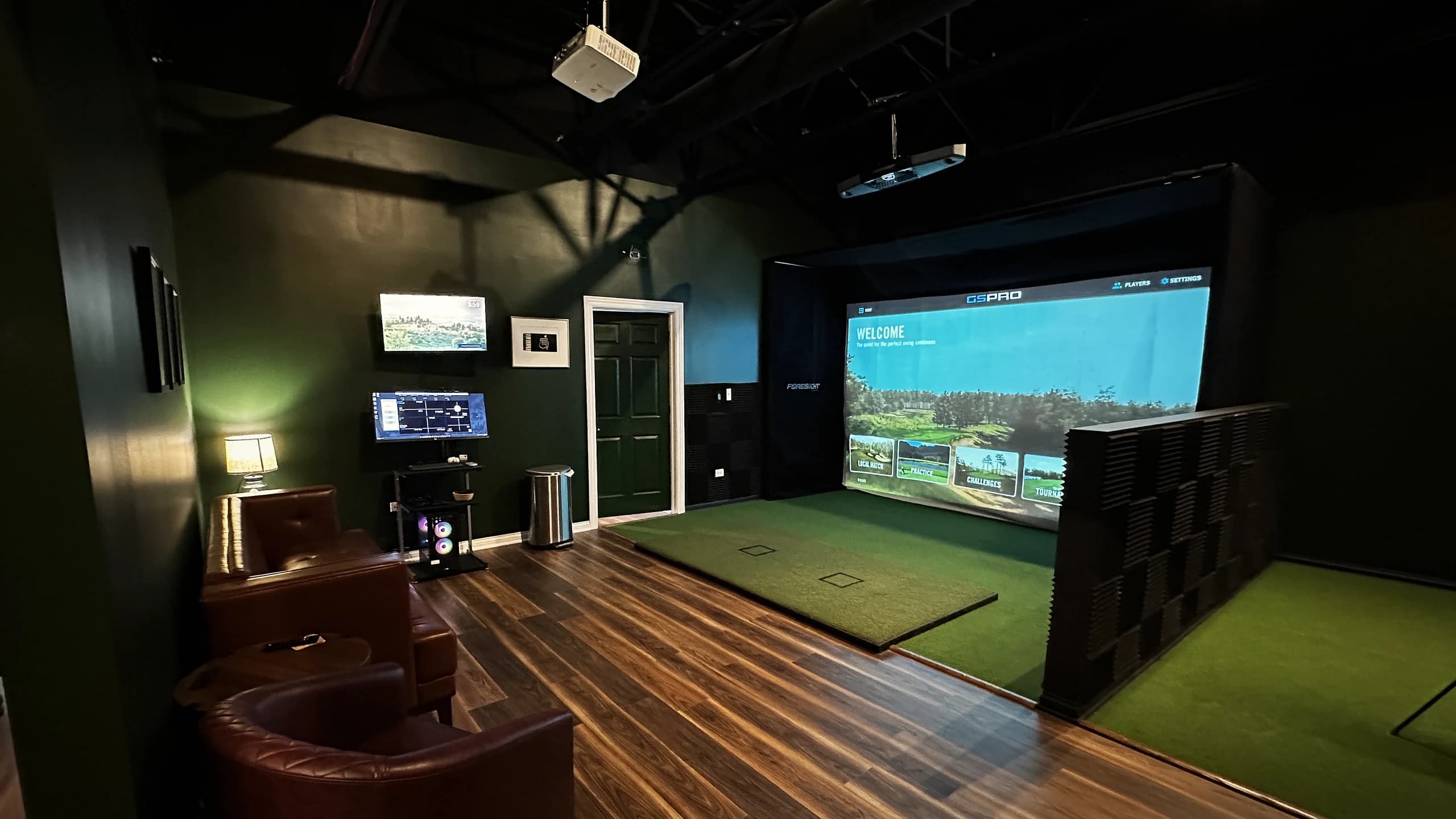 That Golf Place simulator bay with dark walls, warm wood floors, and leather lounge seating