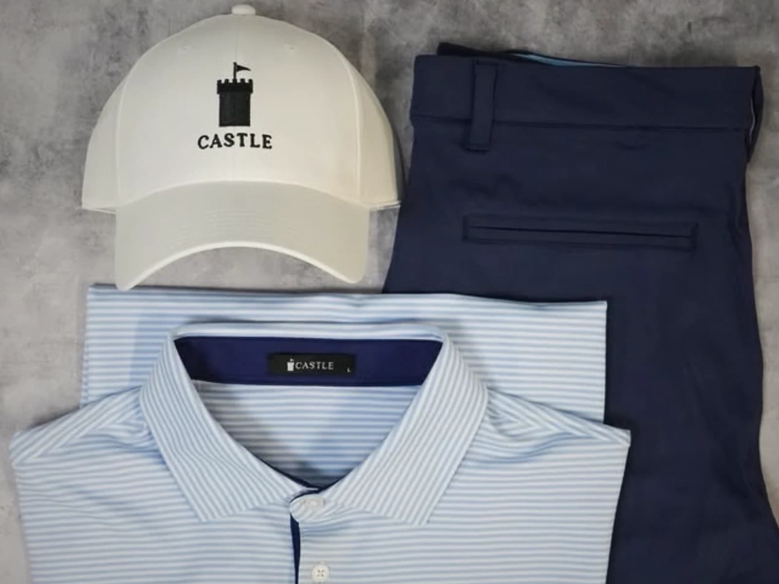Castle Golf Co. flat-lay with white cap with castle pennant logo, navy performance trousers, and light-blue striped polo on a concrete surface.
