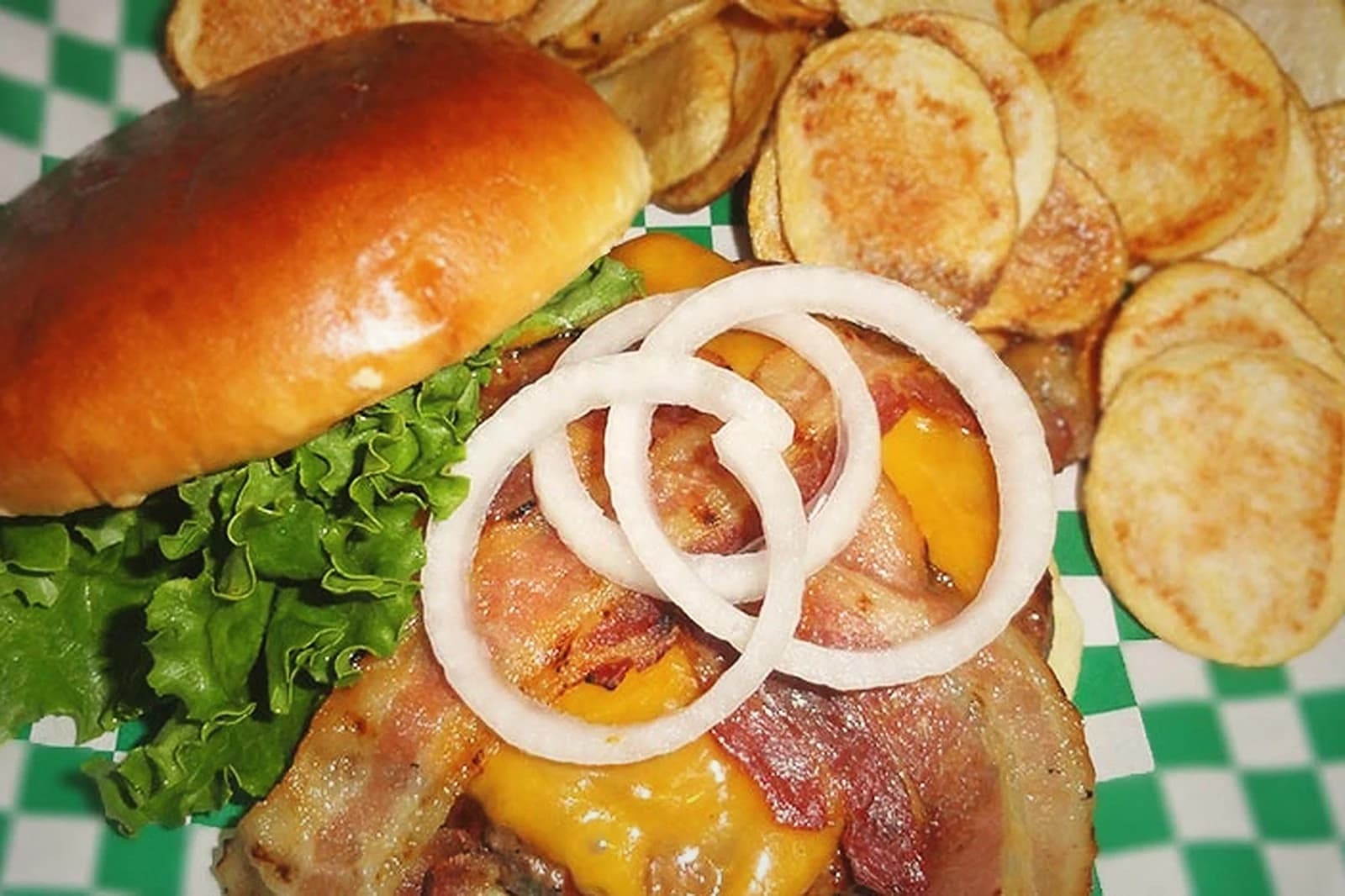 DJ's Sports Bar & Grill bacon cheeseburger with onion rings, lettuce, and a side of housemade chips on green-checkered paper.