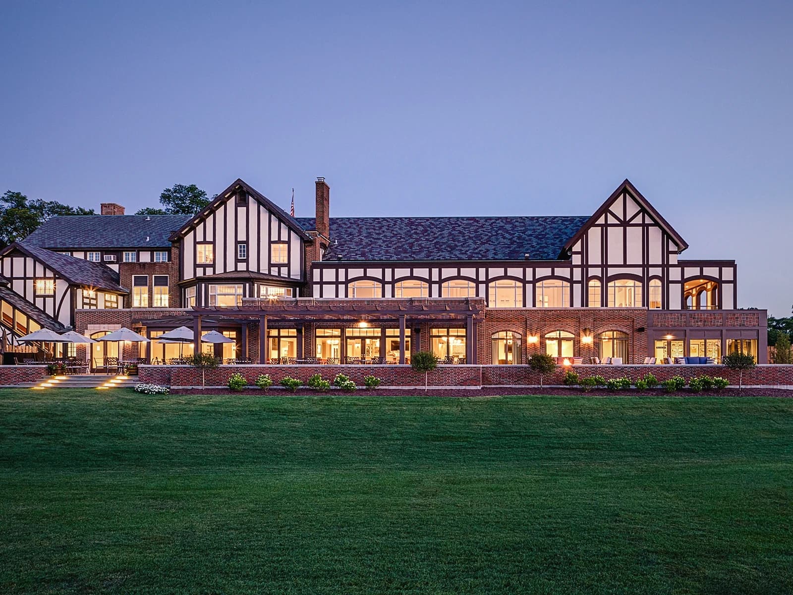 Hinsdale Golf Club Tudor clubhouse at twilight, warm interior lights glowing against a blue evening sky, viewed from the practice lawn.