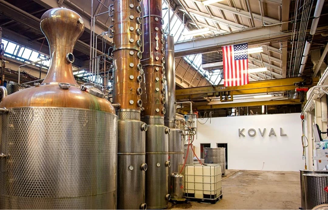 KOVAL Distillery production floor in Chicago: copper-pot stills and stainless fermentation tanks, an American flag hanging from the rafters, and the KOVAL wordmark on the back wall.