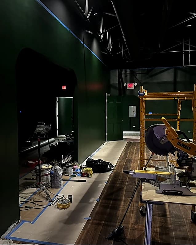 That Golf Place during build-out: forest-green walls, exposed black ceiling structure, hardwood flooring being laid, and a DeWalt miter saw on the construction floor
