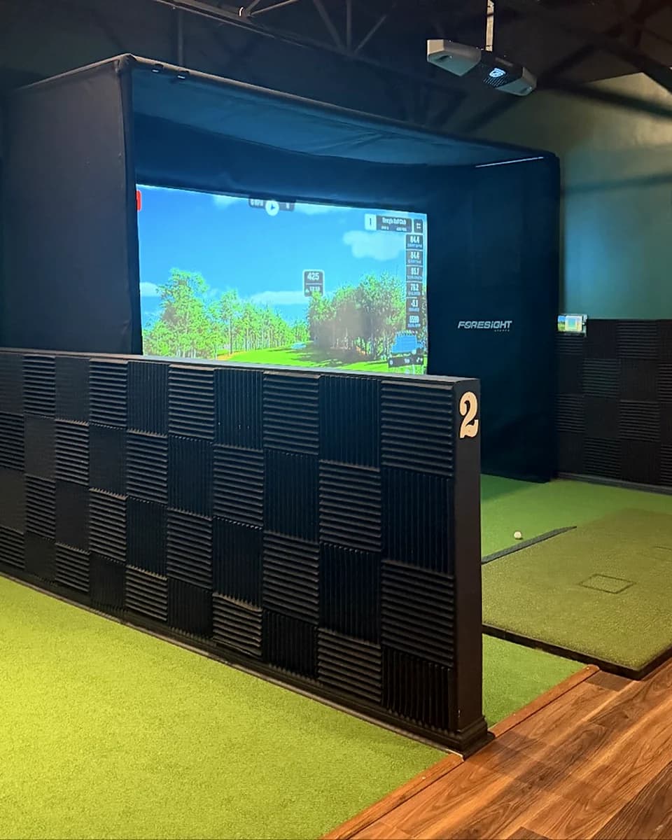 Bay 02 at That Golf Place: an acoustic-foam wedge wall lines the front of the hitting bay, with a Foresight Falcon launch monitor and a lit GSPro course on the impact screen behind — the visual signature of an independently soundproofed simulator bay.
