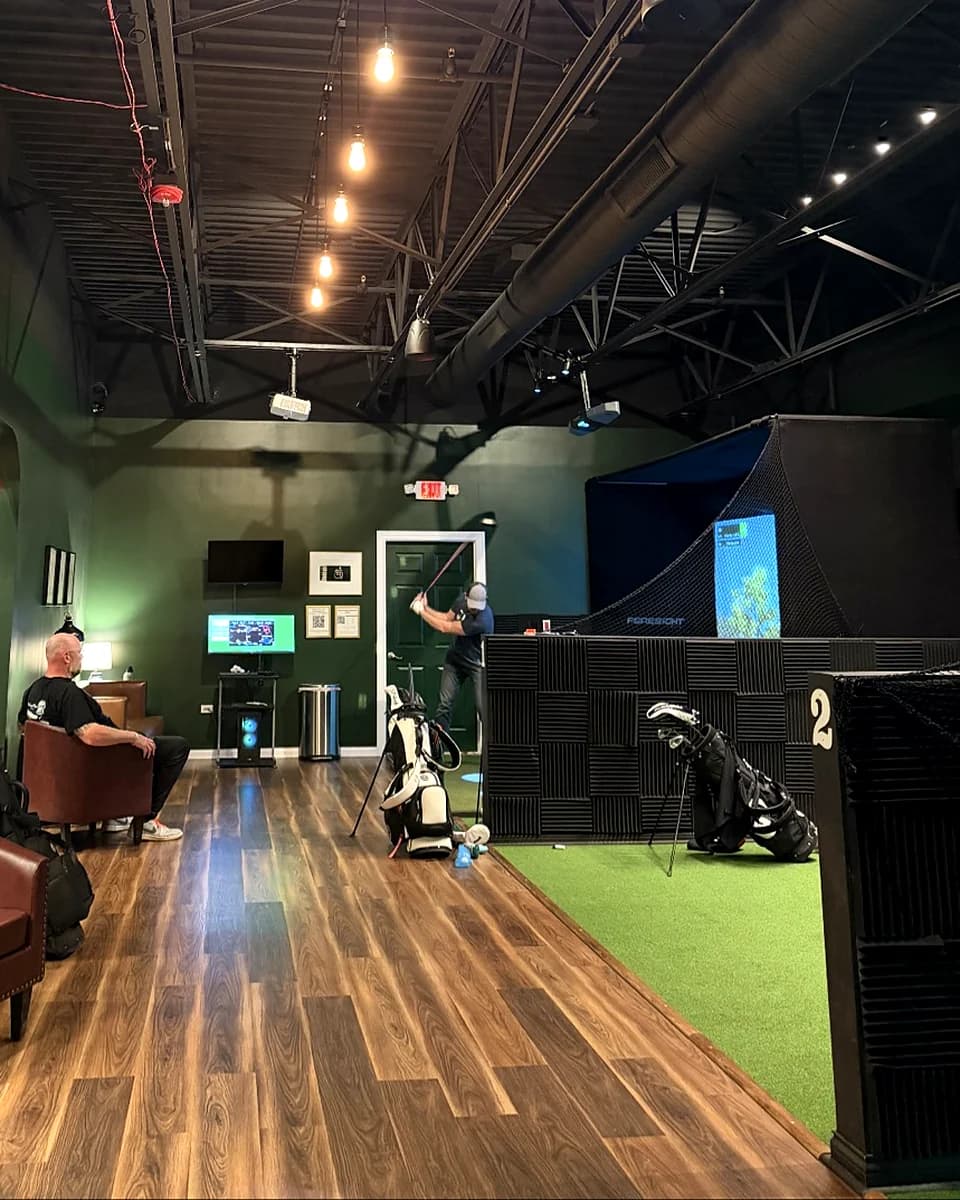 Warm Edison-bulb pendants run across the ceiling at That Golf Place while a member swings in Bay 02 with the Foresight screen lit and no glare — calibrated lighting in practice.