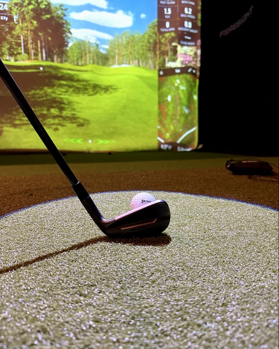 Ground-level view from behind a black iron clubhead at address with a Srixon ball on the premium hitting mat at That Golf Place, the impact screen lit with a tree-lined fairway and Foresight launch monitor data overlaid in the background.