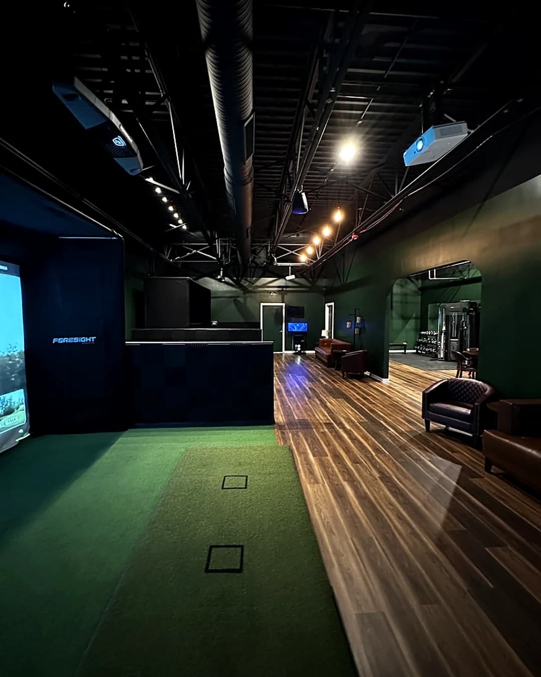 Inside That Golf Place: a soundproofed simulator bay with a Foresight curtain and putting turf in the foreground, leather club chairs in the lounge alongside, and a second bay visible through the open archway.