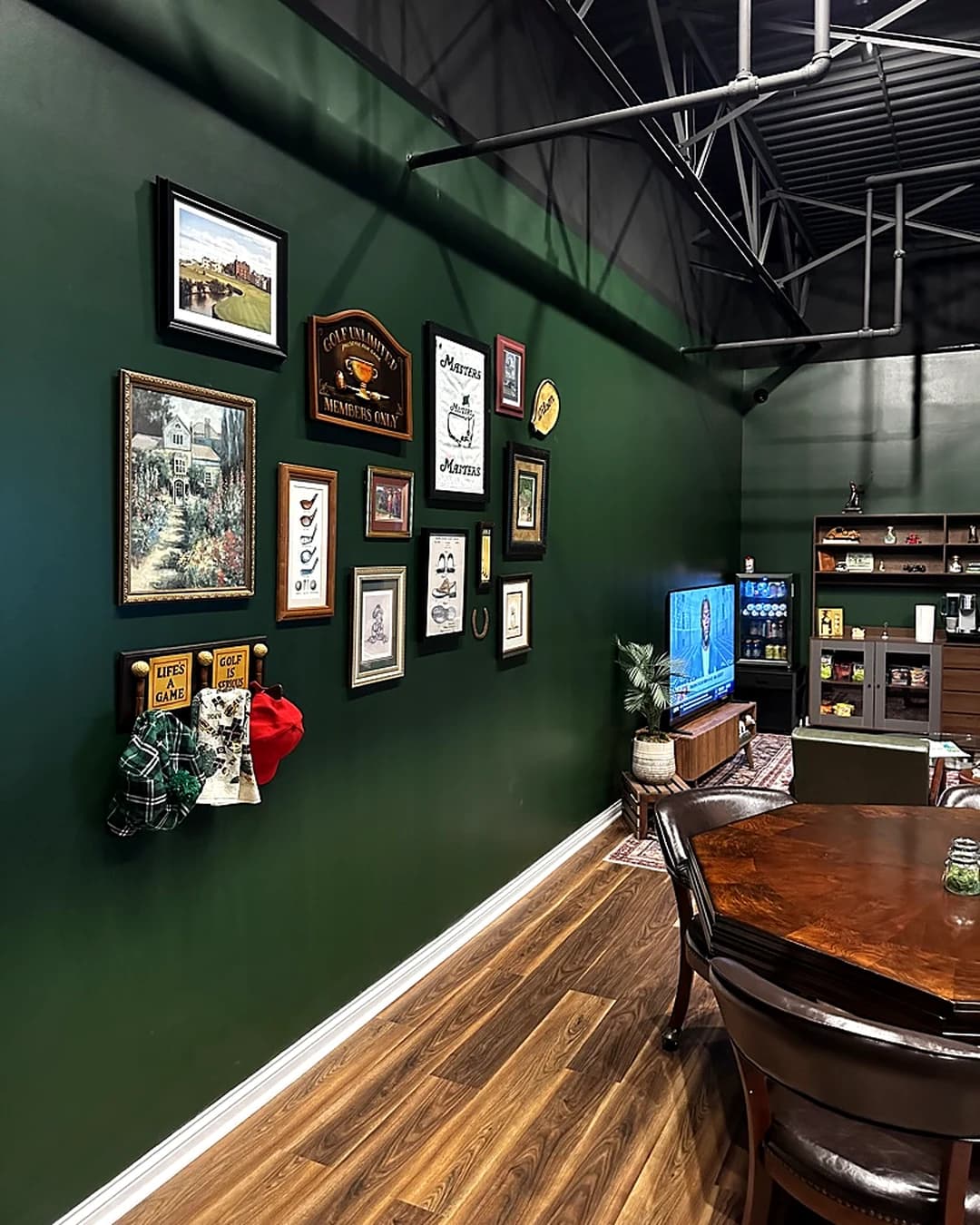 A forest-green clubhouse wall at That Golf Place hung with framed golf course paintings, a Masters tournament print, and vintage "Members Only" signage. A peg rack of plaid and checkered headcovers hangs below. To the right, built-in millwork holds a TV showing tournament coverage, a beverage display, and an espresso setup, with a small dining table in the foreground.