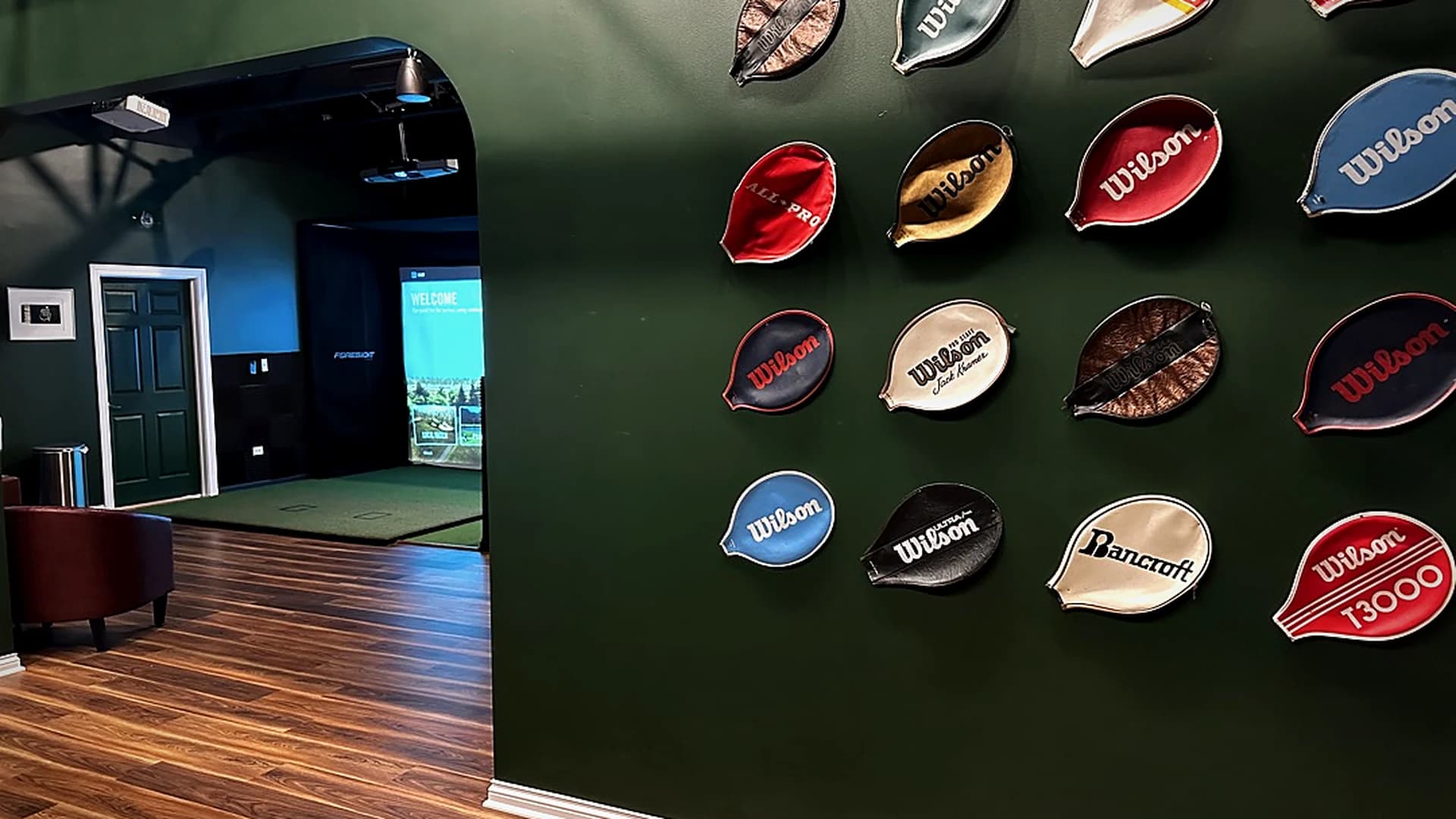 Inside That Golf Place: archway view into a simulator bay with the Foresight WELCOME screen, beside a forest-green wall hung with vintage tennis racket covers.