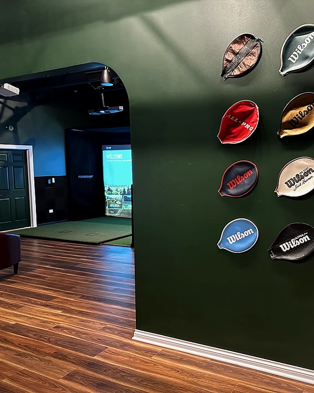 Inside That Golf Place: archway view into a simulator bay with the Foresight WELCOME screen, beside a forest-green wall hung with vintage tennis racket covers.