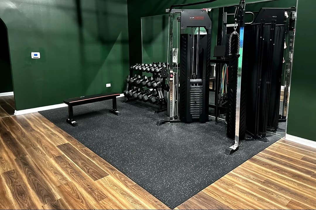 The fitness area at That Golf Place: a forest-green walled room with a black rubber lifting platform on warm wood floors, holding a flat bench, a dumbbell rack, and a CarStrength functional trainer with cable stack and resistance bands.