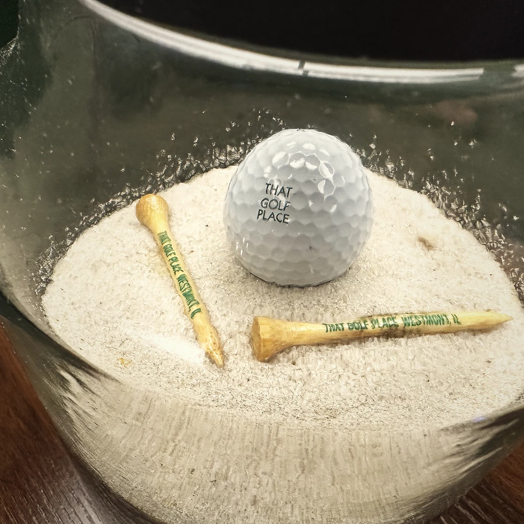 A That Golf Place branded golf ball with the club logo, sitting in a glass cup with two wooden tees printed "That Golf Place, Westmont, IL" on a bed of fine sand.