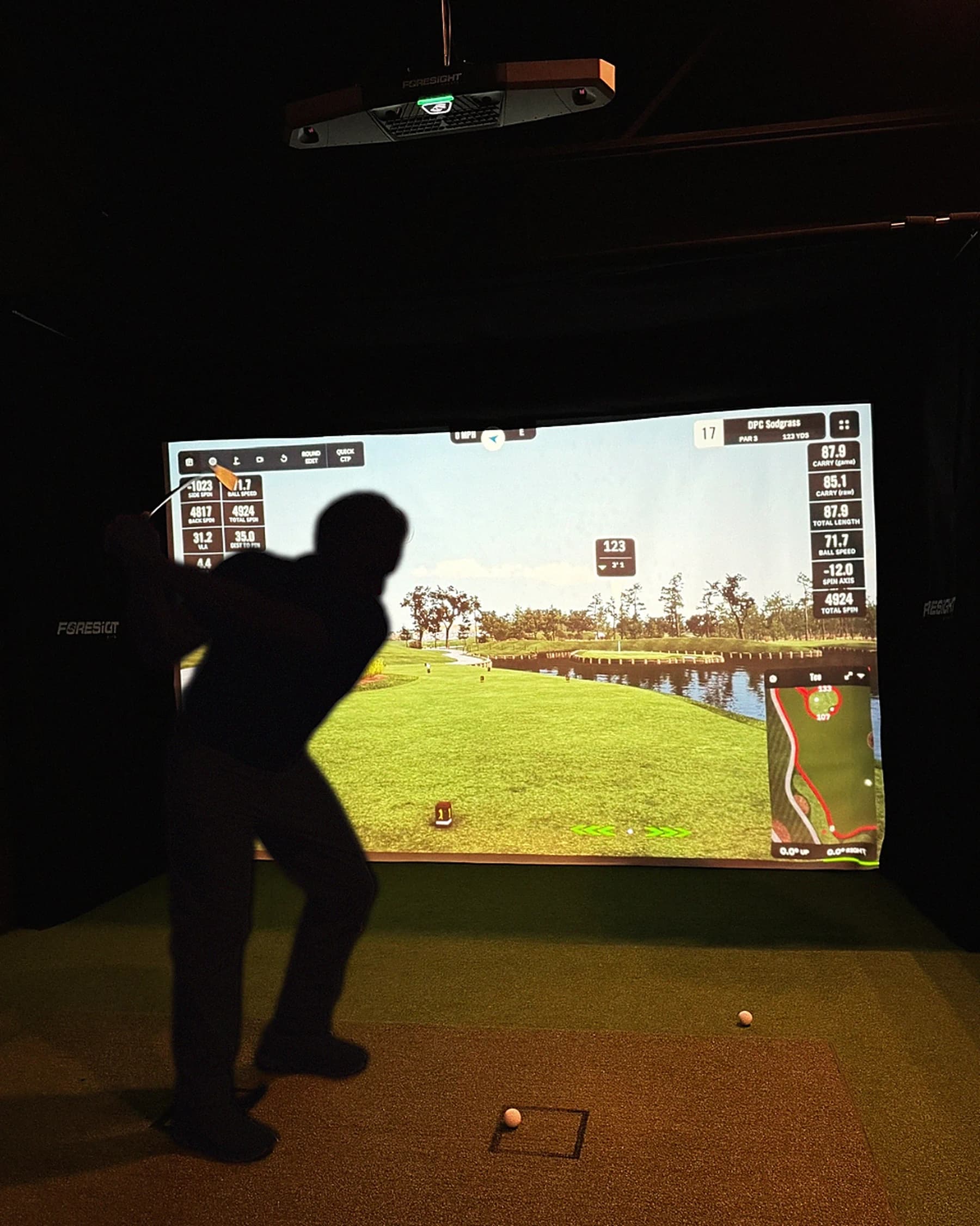 A player in silhouette mid-backswing inside a simulator bay at That Golf Place, the screen displaying a sunlit virtual course with live shot data overlays for ball speed, carry, side spin, and total spin.