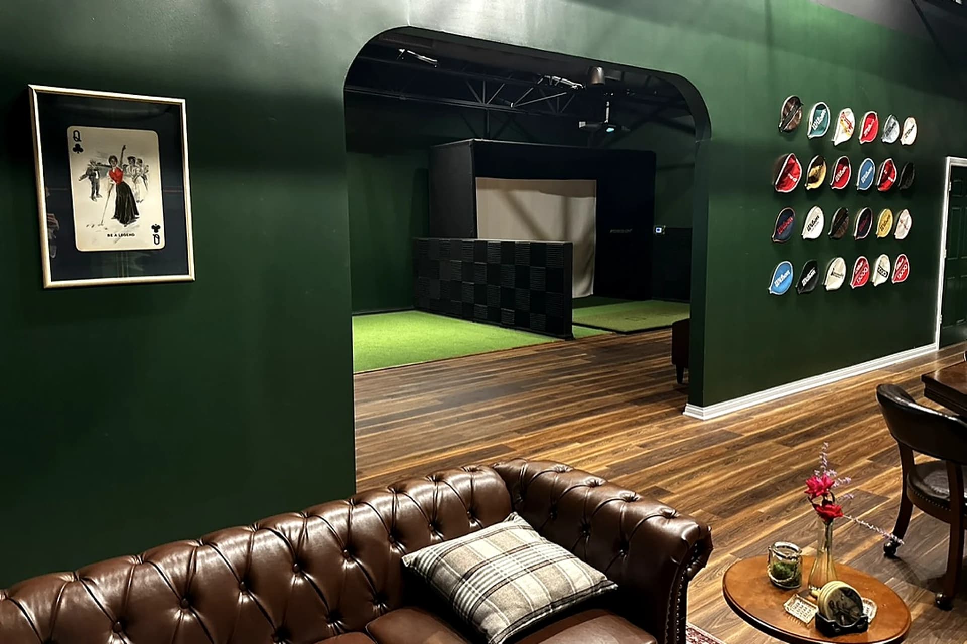 The members lounge at That Golf Place: a tufted brown leather Chesterfield sofa with a tartan throw pillow, a side table with a candle and floral arrangement, a framed "Be a Legend" Queen of Clubs print on the forest-green wall, and an open archway behind revealing a simulator bay with putting turf and impact screen. The right wall holds a continuation of the vintage tennis-racket-cover gallery.